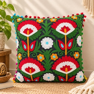 May include: A square throw pillow with a dark navy blue base featuring vibrant, multi-colored floral embroidery in red, white, orange, and green. The edges are decorated with small, colorful pom-poms in red, blue, and yellow.