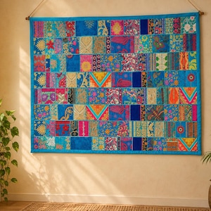 Op de afbeelding: Een rechthoekige wandhanger met een levendig patchworkontwerp bestaande uit talloze kleine stoffen vierkantjes in tinten blauw, roze, oranje en goud, met ingewikkelde bloem- en geometrische patronen, afgewerkt met een effen blauwe rand.