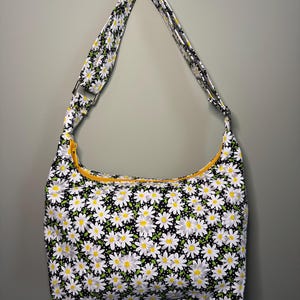 May include: A hobo-style shoulder bag featuring a black fabric base with a white daisy floral pattern, green leaves, and small white polka dots. The bag includes a bright yellow zipper closure along the top and a matching patterned shoulder strap.