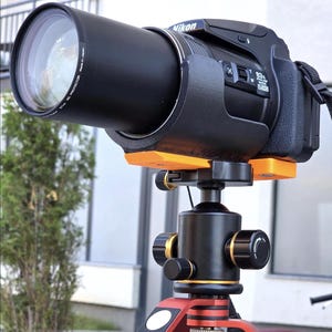 Op de afbeelding: Een zwarte Nikon digitale camera met een 83x optische zoomlens gemonteerd op een statief. De camera is bevestigd aan de statiefkop met een feloranje montagebeugel. De statiefkop is zwart met gouden accenten en de statiefpoten zijn rood.