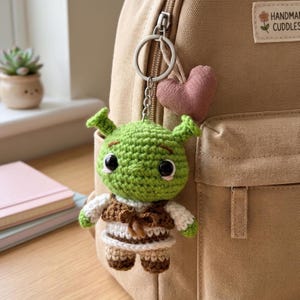 Puede incluir: Un llavero de ganchillo con un personaje de ogro verde de grandes ojos negros, que lleva un chaleco marrón y una camisa blanca, unido a una anilla de metal plateado con un pequeño corazón de tela rosa, colgado de una mochila de lona beige.