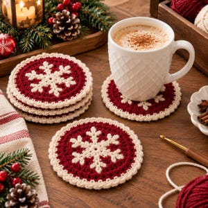 May include: A set of round, crocheted coasters featuring a deep red background with a cream-colored snowflake pattern in the center. The coasters have a cream-colored border and are displayed on a wooden surface alongside a white mug.