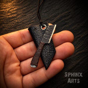 May include: A miniature cleaver pendant featuring a dark, textured metal blade with a brown cord-wrapped handle. The cleaver is attached to a black leather sheath with a silver-toned metal eyelet and a thin black cord for wearing as a necklace.