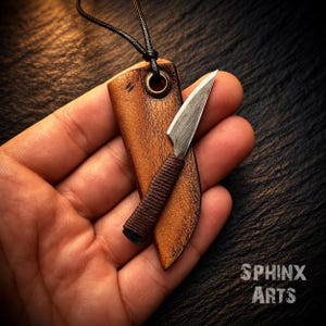 May include: A small metal knife with a dark brown cord-wrapped handle, housed in a tan leather sheath with a metal eyelet and a black cord necklace. The text SPHINX ARTS is visible in the lower right corner of the image, set against a dark background.