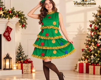 Op de afbeelding: Een mouwloze groene jurk ontworpen als een kerstboom, voorzien van horizontale rijen gouden klatergoudfranjes en ronde rode, blauwe en gele ornamenten. De outfit bevat een bijpassende hoofdband met een kleine boom als versiering.