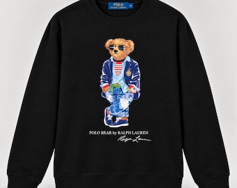 Op de afbeelding: Een zwart sweatshirt met lange mouwen en een afbeelding van een teddybeer met een zonnebril, een marineblauw jasje, een rood-wit gestreept shirt en een blauwe spijkerbroek. De tekst POLO BEAR BY RALPH LAUREN en een handtekening staan eronder.