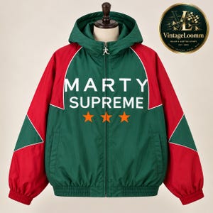 Op de afbeelding: Een groen en rood jack met capuchon en ritssluiting, voorzien van de witte tekst MARTY SUPREME en drie oranje sterren op de voorzijde. Het jack heeft lange mouwen met rode panelen, een volledige ritssluiting met een loper en boorden.