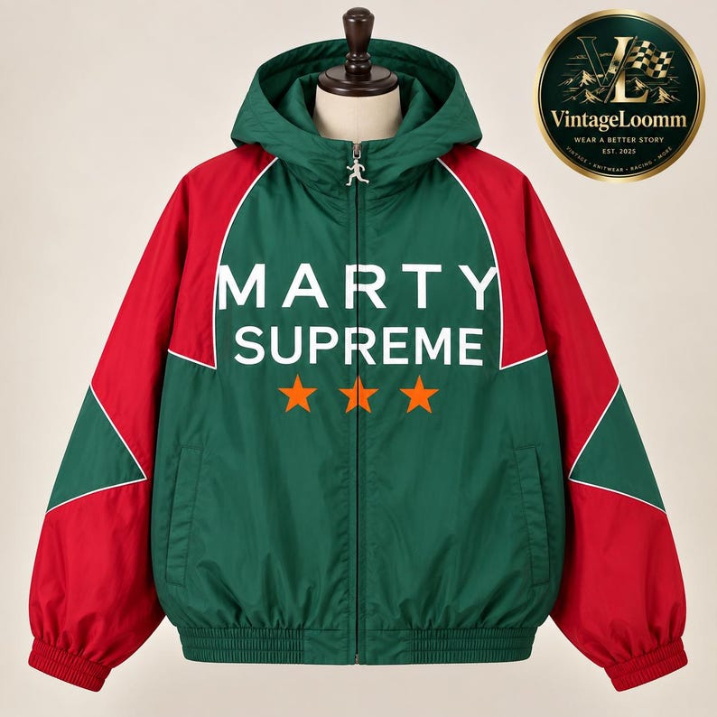 Op de afbeelding: Een groen en rood jack met capuchon en ritssluiting, voorzien van de witte tekst MARTY SUPREME en drie oranje sterren op de voorzijde. Het jack heeft lange mouwen met rode panelen, een volledige ritssluiting met een loper en boorden.