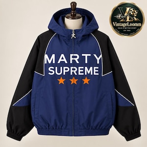 Peut inclure: Un coupe-vent à capuche bleu marine et noir avec le texte blanc MARTY SUPREME et trois étoiles orange sur la poitrine. La veste possède une fermeture éclair intégrale sur le devant avec une tirette argentée, des manches longues et une capuche.