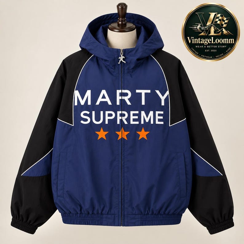 Op de afbeelding: Een marineblauw en zwart windjack met capuchon, voorzien van de witte tekst MARTY SUPREME en drie oranje sterren op de borst. Het jack heeft een volledige ritssluiting aan de voorzijde met een zilverkleurige trekker, lange mouwen en een capuchon.