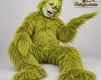Può includere: Un costume integrale in pelliccia sintetica verde con una maschera di personaggio dettagliata con naso a punta, fronte corrugata e bocca aperta. La tuta è ricoperta di lunghe fibre verdi arruffate. Il logo VintageLoomm è visibile nell'angolo.