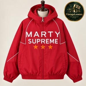 Op de afbeelding: Een felrood jack met capuchon en ritssluiting, voorzien van witte biezen langs de schouders en borst. Op de voorzijde staat de witte tekst MARTY SUPREME boven drie oranje sterren. Een kleine zilverkleurige hanger zit aan de ritssluiting.