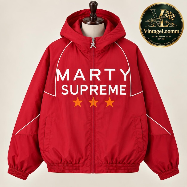 Op de afbeelding: Een felrood jack met capuchon en ritssluiting, voorzien van witte biezen langs de schouders en borst. Op de voorzijde staat de witte tekst MARTY SUPREME boven drie oranje sterren. Een kleine zilverkleurige hanger zit aan de ritssluiting.
