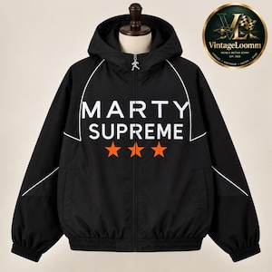 Op de afbeelding: Een zwarte jas met capuchon en ritssluiting, voorzien van witte biezen langs de schouders en borst. Op de voorzijde staat de witte tekst MARTY SUPREME boven drie oranje sterren. Een zilveren ritstrekker in de vorm van een hardloper is bevestigd.