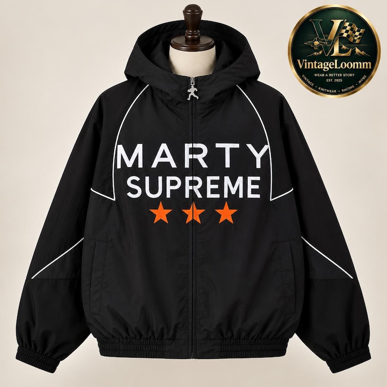 Op de afbeelding: Een zwarte jas met capuchon en ritssluiting, voorzien van witte biezen langs de schouders en borst. Op de voorzijde staat de witte tekst MARTY SUPREME boven drie oranje sterren. Een zilveren ritstrekker in de vorm van een hardloper is bevestigd.