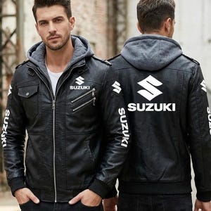 May include: A black faux leather jacket featuring a grey hooded insert, a front zip fastening, and multiple pockets. The Suzuki logo and brand name are printed in white on the chest, sleeves, and back. The jacket has ribbed cuffs and hem.
