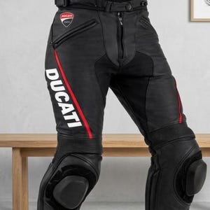 Może przedstawiać: Czarne skórzane spodnie motocyklowe z białym logo Ducati na prawym udzie, czerwonym pionowym paskiem, kieszeniami na zamek błyskawiczny, paskiem w talii i zintegrowanymi czarnymi ochraniaczami kolan. Elastyczne panele nad kolanami.