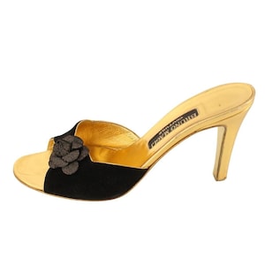 Op de afbeelding: Goudkleurige muiltjes met hoge hakken, met een zwarte suède neus en een decoratieve bloem. De schoen heeft een gouden binnenzool en hak. De merknaam "BRUNO MAGLI COUTURE" staat op de binnenzool.