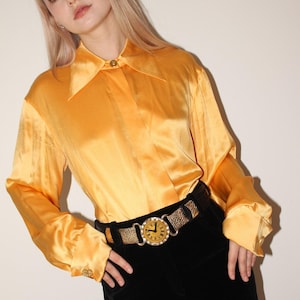May include: A golden yellow satin blouse with a pointed collar and buttoned cuffs. The blouse is paired with black velvet pants and a gold and leopard print belt with a clock-face buckle.