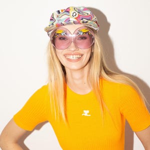 Puede incluir: Una mujer con una gorra estampada colorida y gafas de sol rosas grandes. Lleva una camiseta de punto acanalado naranja brillante de manga corta con un peque&ntilde;o logotipo blanco. El fondo es blanco liso.