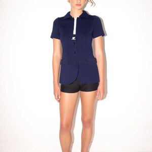 K&ouml;nnte beinhalten: Marineblaue Jacke mit kurzen &Auml;rmeln und wei&szlig;em Streifen vorne, kombiniert mit schwarzen Shorts. Die Jacke hat zwei Vordertaschen und wird mit schwarzen Abs&auml;tzen getragen. Das Outfit ist vor einem wei&szlig;en Hintergrund abgebildet.