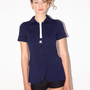 K&ouml;nnte beinhalten: Marineblaue Jacke mit kurzen &Auml;rmeln, wei&szlig;em Rei&szlig;verschluss und Kragen. Die Jacke hat zwei Vordertaschen und schwarze Kn&ouml;pfe. Das Model tr&auml;gt schwarze Shorts. Die Jacke ist im Vintage-Stil.
