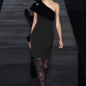 May include: A woman wearing a black one-shoulder dress with a black velvet overlay and black lace tights. She is walking on a runway.