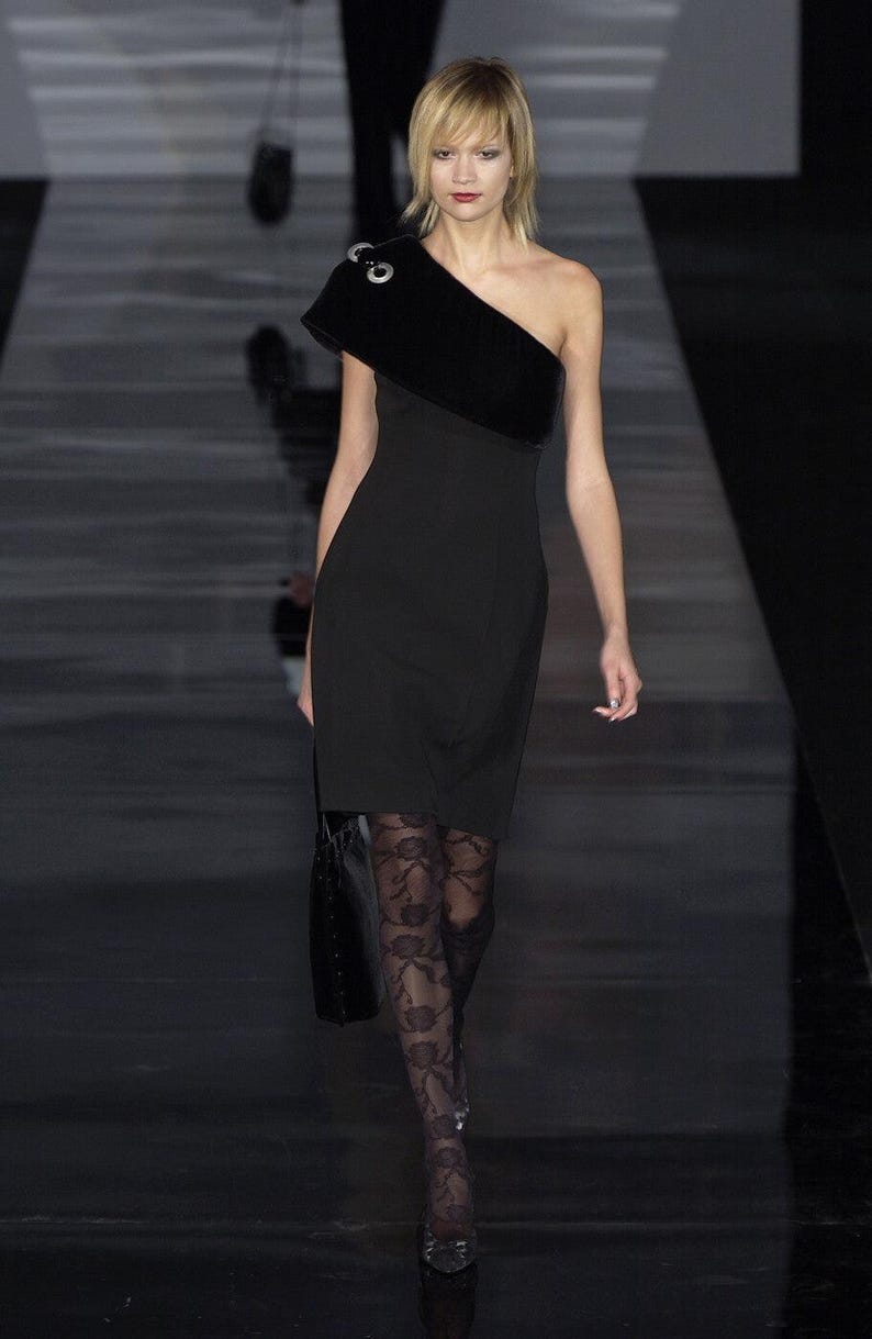 May include: A woman wearing a black one-shoulder dress with a black velvet overlay and black lace tights. She is walking on a runway.