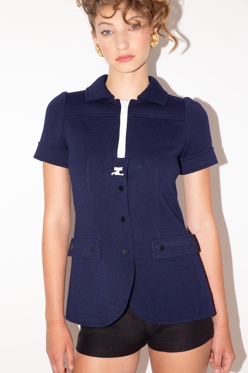 K&ouml;nnte beinhalten: Marineblaue Jacke mit kurzen &Auml;rmeln, wei&szlig;em Rei&szlig;verschluss und Kragen. Die Jacke hat zwei Vordertaschen und schwarze Kn&ouml;pfe. Ein Kleidungsst&uuml;ck im Vintage-Stil.