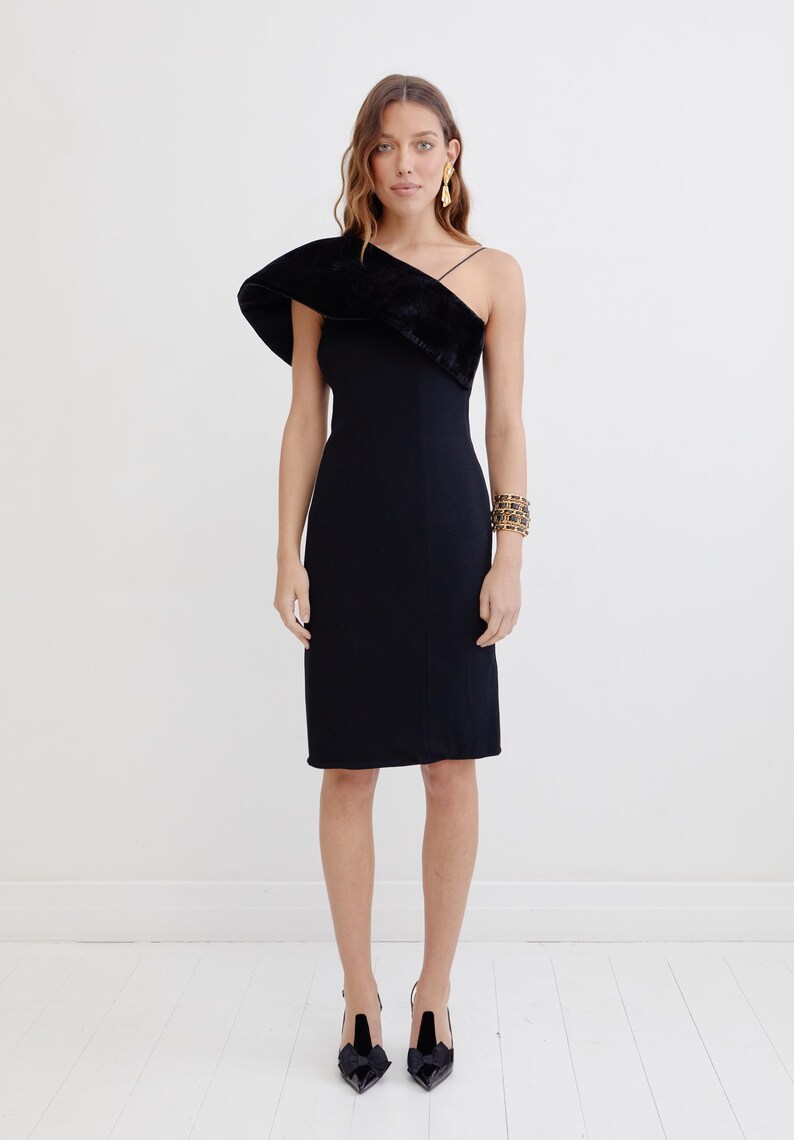 May include: A black one-shoulder dress with a velvet trim detail. The dress has a fitted silhouette and a midi length. The model is wearing black heels.