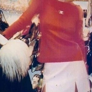 Puede incluir: Una mujer sonr&iacute;e, vestida con un top rojo de manga larga y una minifalda blanca con una abertura. El fondo muestra zapatos y otros art&iacute;culos.