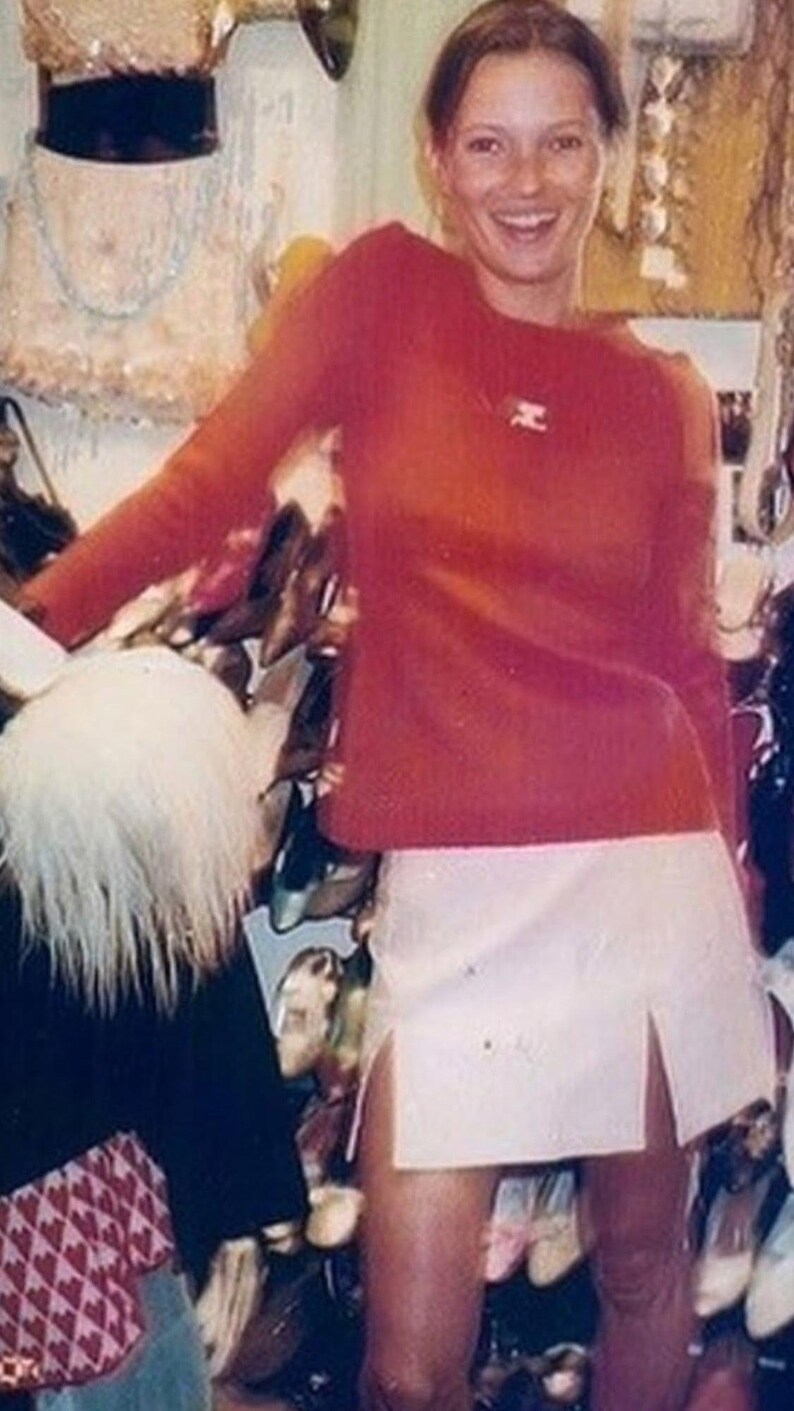 Puede incluir: Una mujer sonr&iacute;e, vestida con un top rojo de manga larga y una minifalda blanca con una abertura. El fondo muestra zapatos y otros art&iacute;culos.