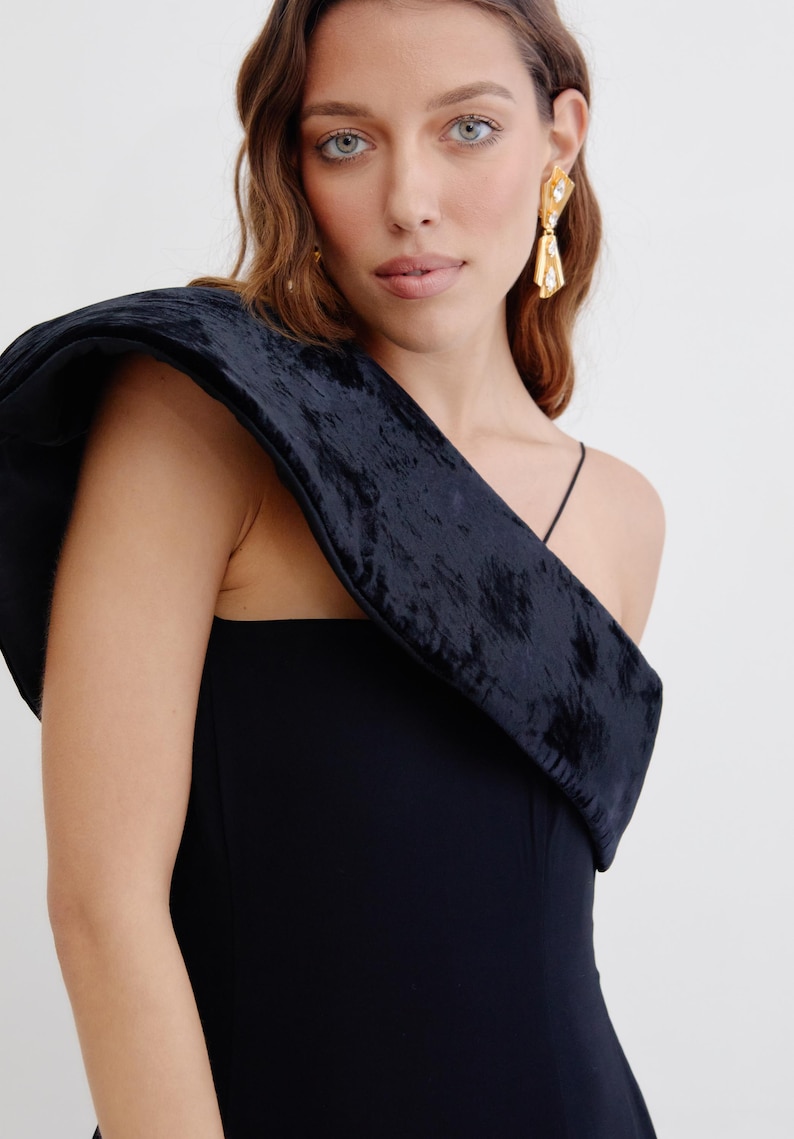 May include: A black one-shoulder dress with a velvet detail on the shoulder. The dress has a fitted silhouette and a simple design.