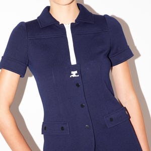 K&ouml;nnte beinhalten: Marineblaue Jacke mit kurzen &Auml;rmeln, wei&szlig;em Rei&szlig;verschluss und kleinem wei&szlig;em Emblem. Sie hat einen Kragen, schwarze Kn&ouml;pfe und zwei Vordertaschen. Die &Auml;rmel sind umgeschlagen.