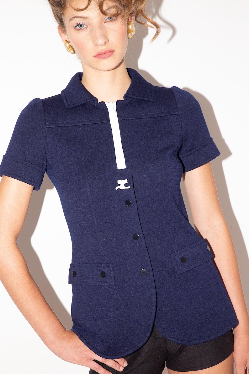 K&ouml;nnte beinhalten: Marineblaue Jacke mit kurzen &Auml;rmeln, wei&szlig;em Rei&szlig;verschluss und kleinem wei&szlig;em Emblem. Sie hat einen Kragen, schwarze Kn&ouml;pfe und zwei Vordertaschen. Die &Auml;rmel sind umgeschlagen.