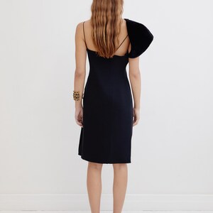 May include: A black sleeveless dress with a single velvet puff sleeve on the left shoulder. The dress has a fitted bodice and a straight skirt.