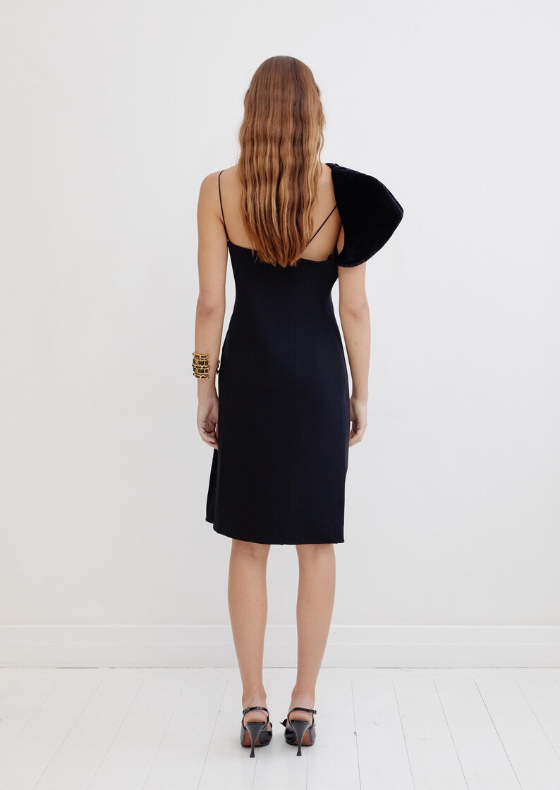May include: A black sleeveless dress with a single velvet puff sleeve on the left shoulder. The dress has a fitted bodice and a straight skirt.