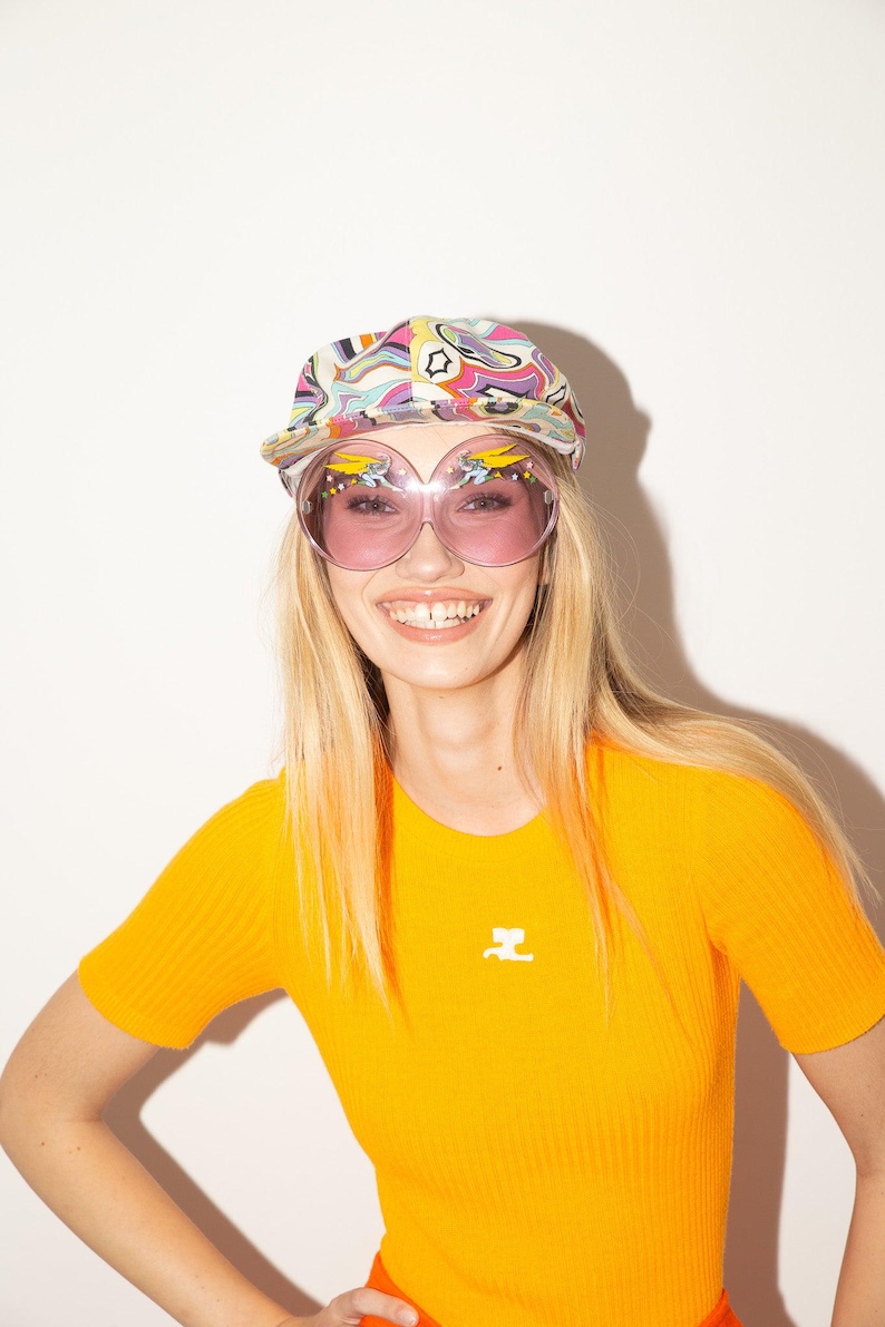 Puede incluir: Una mujer con una gorra estampada colorida y gafas de sol rosas grandes. Lleva una camiseta de punto acanalado naranja brillante de manga corta con un peque&ntilde;o logotipo blanco. El fondo es blanco liso.