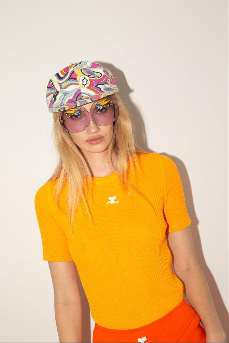 Puede incluir: Una mujer con una camiseta naranja brillante de manga corta y canal&eacute; con un peque&ntilde;o emblema blanco. Tambi&eacute;n lleva una gorra estampada colorida y gafas de sol rosas grandes. El conjunto se completa con una parte inferior naranja.