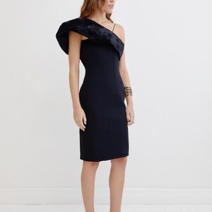 May include: A black one-shoulder dress with a velvet detail on the shoulder. The dress is fitted and has a midi length. The model is wearing black heels with a bow detail.