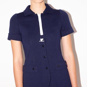 K&ouml;nnte beinhalten: Marineblaue Jacke mit kurzen &Auml;rmeln, wei&szlig;em Rei&szlig;verschluss und Kragen. Die Jacke hat schwarze Kn&ouml;pfe und zwei Vordertaschen. Das Model tr&auml;gt goldene Ohrringe.