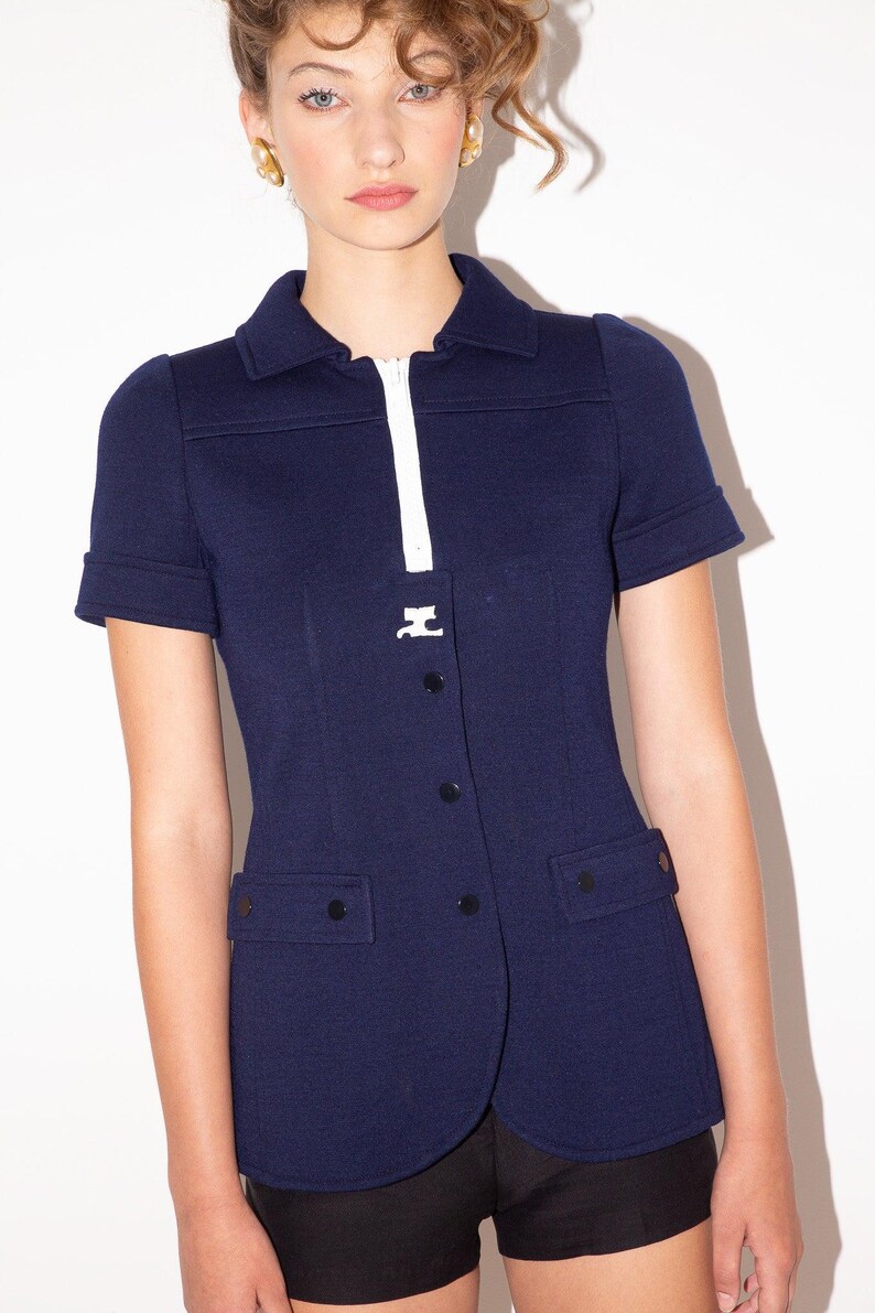 K&ouml;nnte beinhalten: Marineblaue Jacke mit kurzen &Auml;rmeln, wei&szlig;em Rei&szlig;verschluss und Kragen. Die Jacke hat schwarze Kn&ouml;pfe und zwei Vordertaschen. Das Model tr&auml;gt goldene Ohrringe.