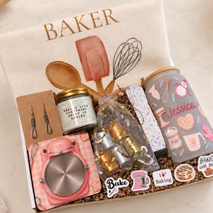 May include: A gift box containing a white towel with the word BAKER and kitchen tools, a pair of whisk earrings, a candle, a frosted glass tumbler with the name Jessica, a pink stand mixer coaster, metallic charms, and baking stickers.