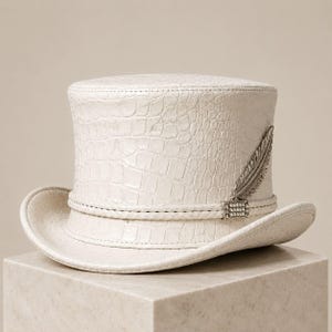 May include: A white top hat featuring a textured crocodile skin pattern. The hat is adorned with a braided band around the base and a silver-toned metallic feather accent embellished with small clear crystals at the center of the band.
