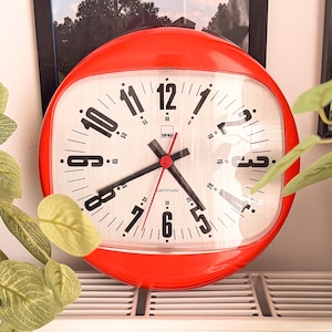 Puede incluir: Un reloj de pared redondo con un marco rojo brillante y una esfera blanca. El reloj presenta números arábigos negros, manecillas de hora y minuto negras y un segundero rojo fino. El texto "isba" y "automatic" está impreso en negro en la esfera.