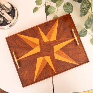 Op de afbeelding: Een rechthoekig houten dienblad met een geometrisch sterpatroon gemaakt van contrasterende donkerbruine en lichtbruine houtinleg. Het dienblad is voorzien van twee gepolijste goudkleurige metalen handgrepen aan de zijkanten voor gemakkelijk dragen.