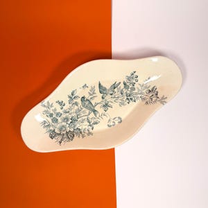 May include: An oval-shaped ceramic dish with an off-white base color, featuring a dark green transferware print of two birds perched on a flowering branch with leaves and berries, centered on the interior surface of the dish.