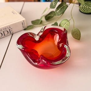 May include: A bowl-shaped glass ashtray featuring a vibrant gradient of deep red at the outer edges transitioning into a bright orange centre. The thick glass has a smooth, undulating rim with three distinct indentations for resting items.
