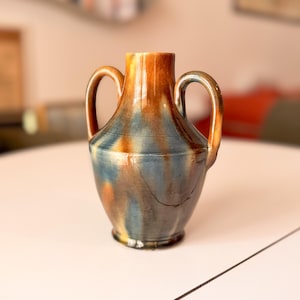 Puede incluir: Un jarrón de cerámica con dos asas, que presenta un esmalte brillante en tonos mezclados de ámbar, marrón y azul pizarra. El recipiente tiene un cuerpo redondeado, un cuello estrecho y un borde ensanchado, con patrones de goteo verticales.
