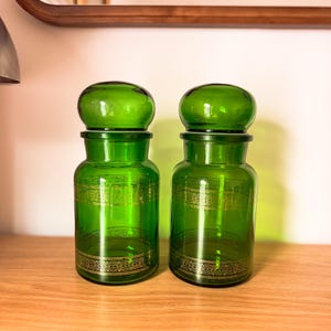 May include: Two matching emerald green glass apothecary jars with rounded lids. Each jar features a cylindrical body adorned with a horizontal band of intricate gold filigree detailing encircling the center and the base of the glass vessel.